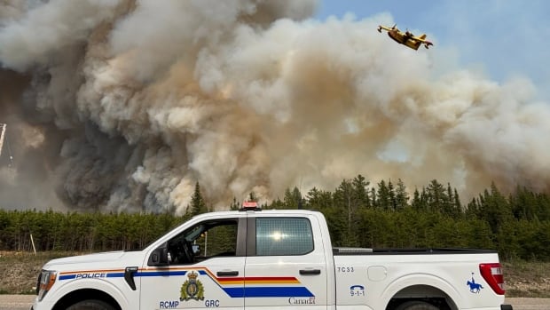Smoke billows into the air in the background with a pickup truck with RCMP marking in the foreground. A yellow airplane flies overhead.