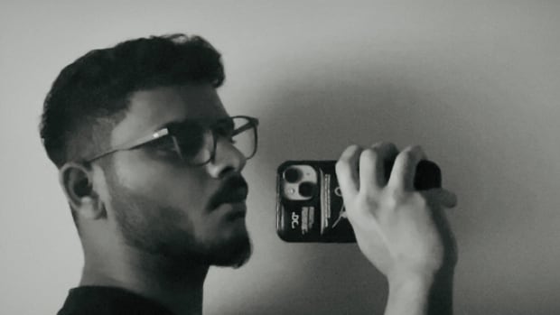 A black and white photo of a man with black hair and dark glasses, holding a cell phone by the side of his face to take a selfie.