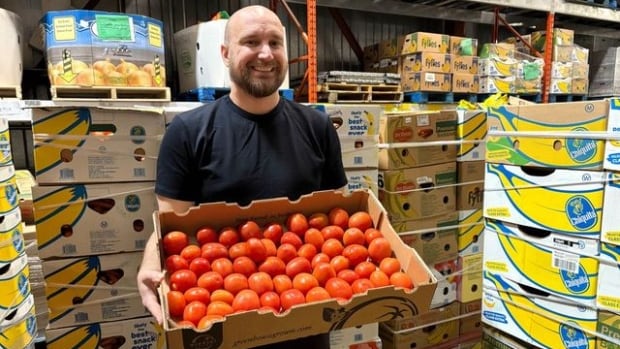 A smiling man holds a large flat box of tomatoes in a warehouse